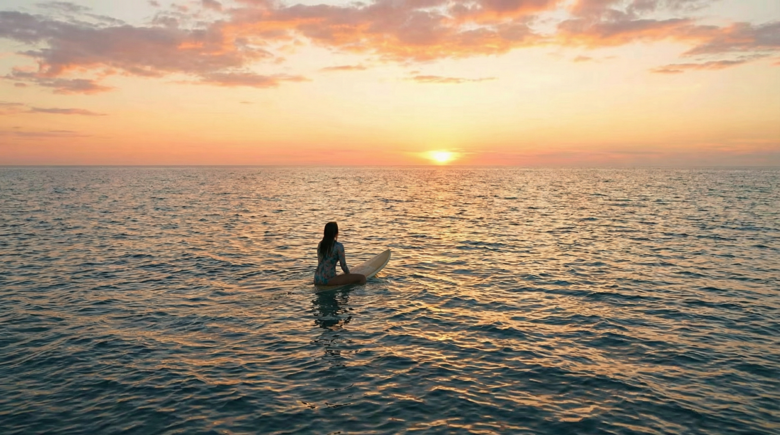 Eine Surferin sitzt ruhig auf einem Surfboard im offenen Meer und blickt bei Sonnenuntergang zum Horizont.